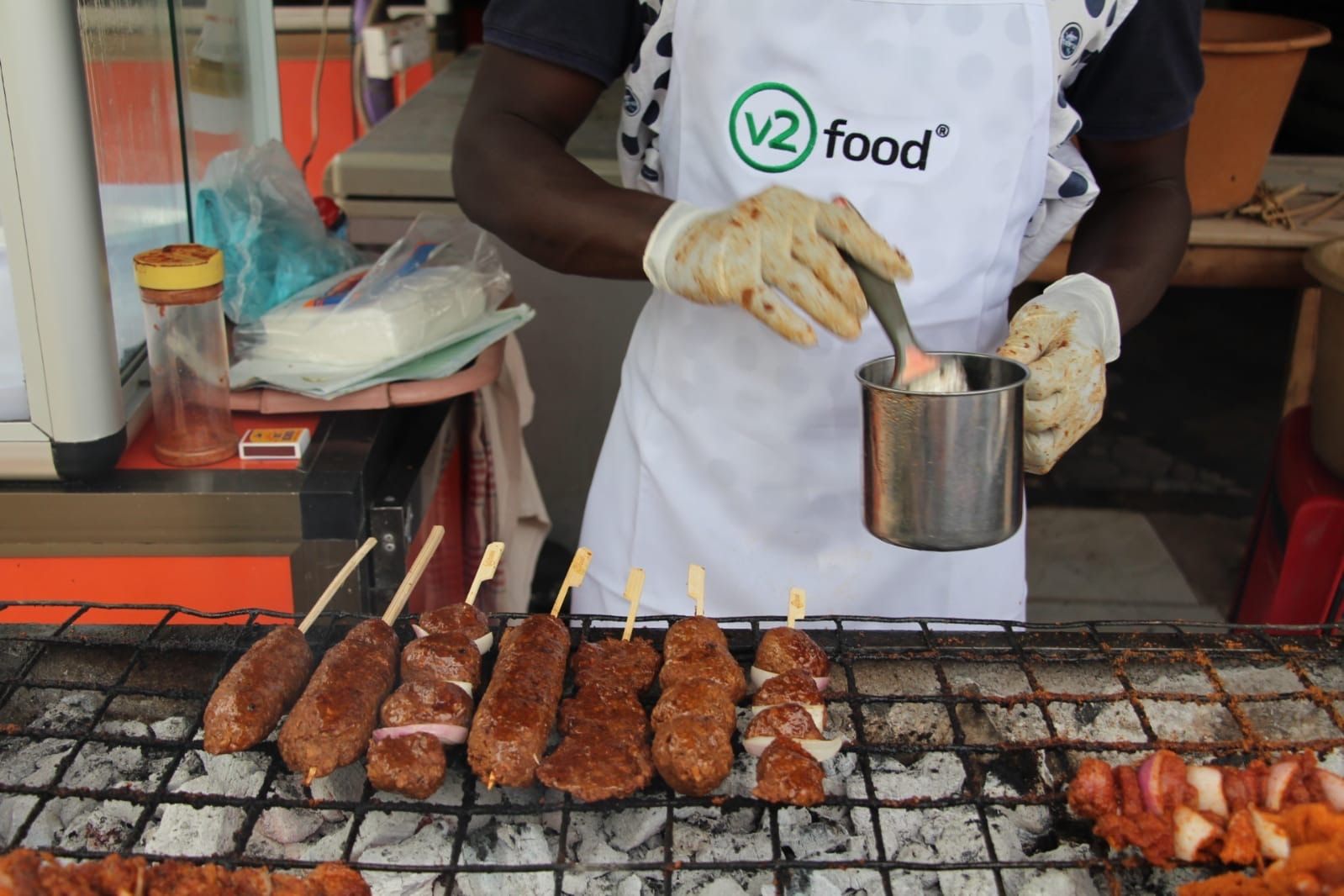 A vendor in a v2food apron grilling skewers at a street stall.